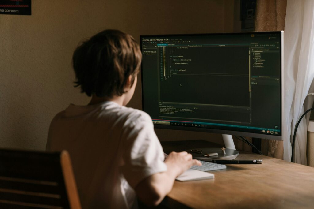 pexels-photo-4709289-4709289 A teenager focused on coding software on a desktop monitor in a home office setting.